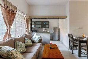 Ein modernes Wohnzimmer mit einer Couch, einem Holztisch und einem Essbereich mit Tisch und Stühlen. Im Hintergrund ist eine Küche mit Geräten und einer Arbeitsfläche erkennbar.