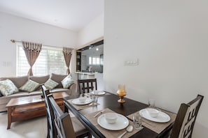 A dining area with a wooden table set for four, a sofa with cushions, and a kitchen in the background. - The Galleria Home by Red Olive Yelahanka (Bengaluru)