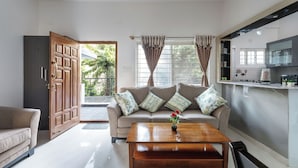 A living room with a sofa, a coffee table with a flower vase, a kitchen area with cabinets, and a large wooden door. - The Galleria Home by Red Olive Yelahanka (Bengaluru)