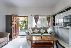 A living room with a sofa, a coffee table with a flower vase, a kitchen area with cabinets, and a large wooden door.