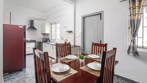 A dining area with a wooden table set for four, surrounded by chairs. There is a kitchen in the background with white cabinets and a red refrigerator. - The Nebula by Red Olive (Bengaluru)