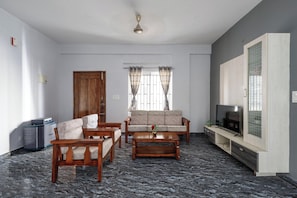 A living room with a sofa, two armchairs, a coffee table, a TV on a wooden stand, and a window with curtains. - The Nebula by Red Olive (Bengaluru)