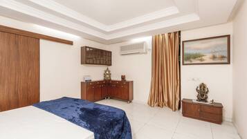 A bedroom with a bed, a wooden wardrobe, a small cabinet, a painting, and an air conditioning unit.