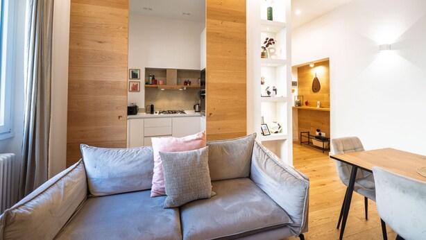 A modern living room with a grey sofa, a wooden accent wall, and a dining area with a wooden table and chairs. - Barbra Suite Testaccio (Roma)