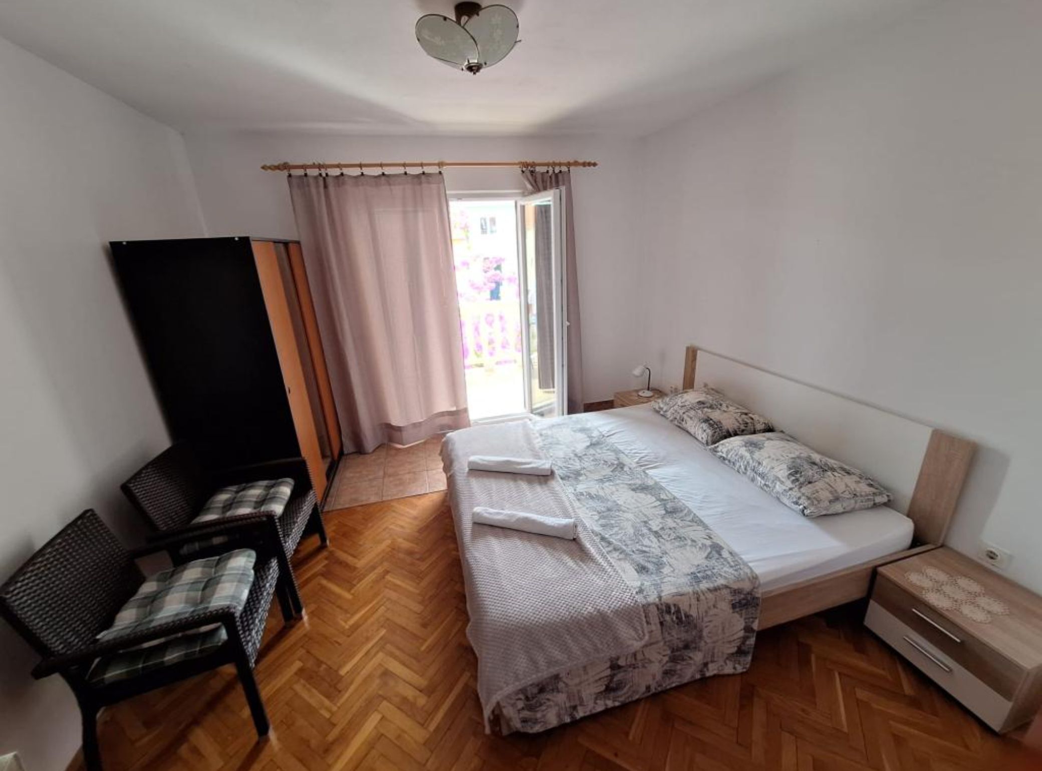 A bedroom with a bed, a chair, a nightstand, a wardrobe, and a window with curtains.