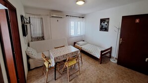 Una habitación pequeña y ordenada con un sofá, una cama individual, una mesa con sillas, un ventanal con persianas, un equipo de aire acondicionado y una puerta con el número 4.