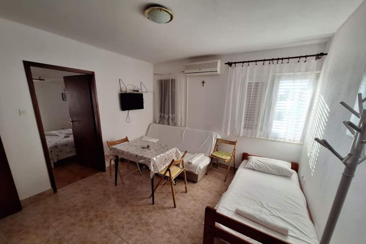 A small, clean room with a bed, a table, and a chair. There's a TV on the wall and an air conditioning unit.