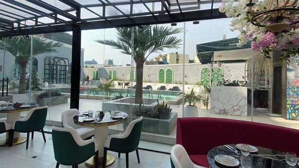 A restaurant with a glass ceiling, outdoor seating, and a view of a pool and buildings. - The Breeze Apartment (Lagos)