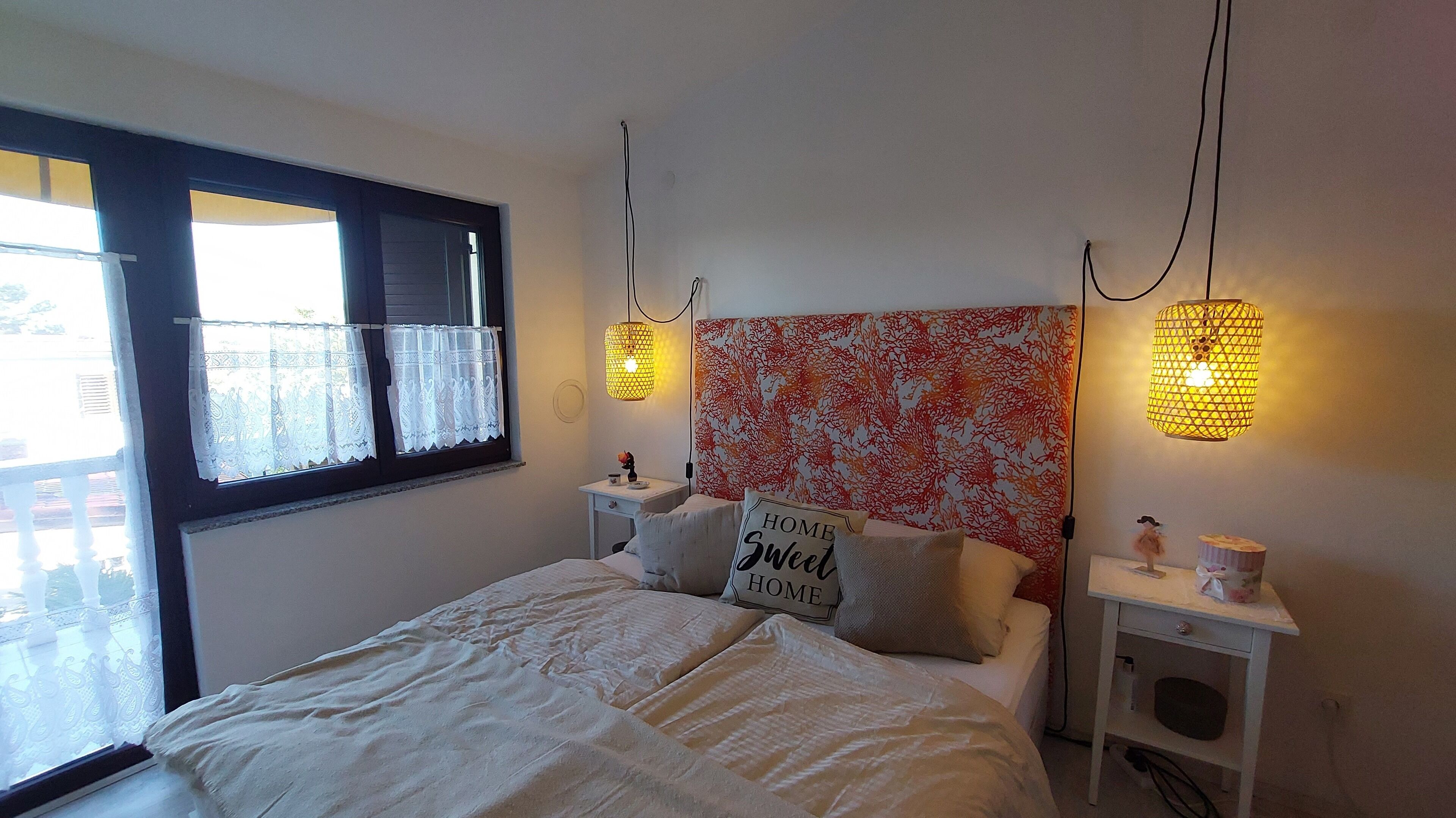 A bedroom with a bed, two bedside tables, a window with lace curtains, and two wall-mounted lamps.