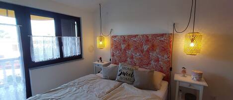 A bedroom with a bed, two bedside tables, a window with lace curtains, and two wall-mounted lamps.