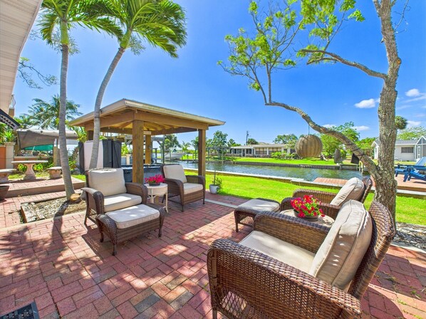 Relax in a serene patio area with a soothing view of the water, surrounded by lush greenery and comfortable wicker furniture.