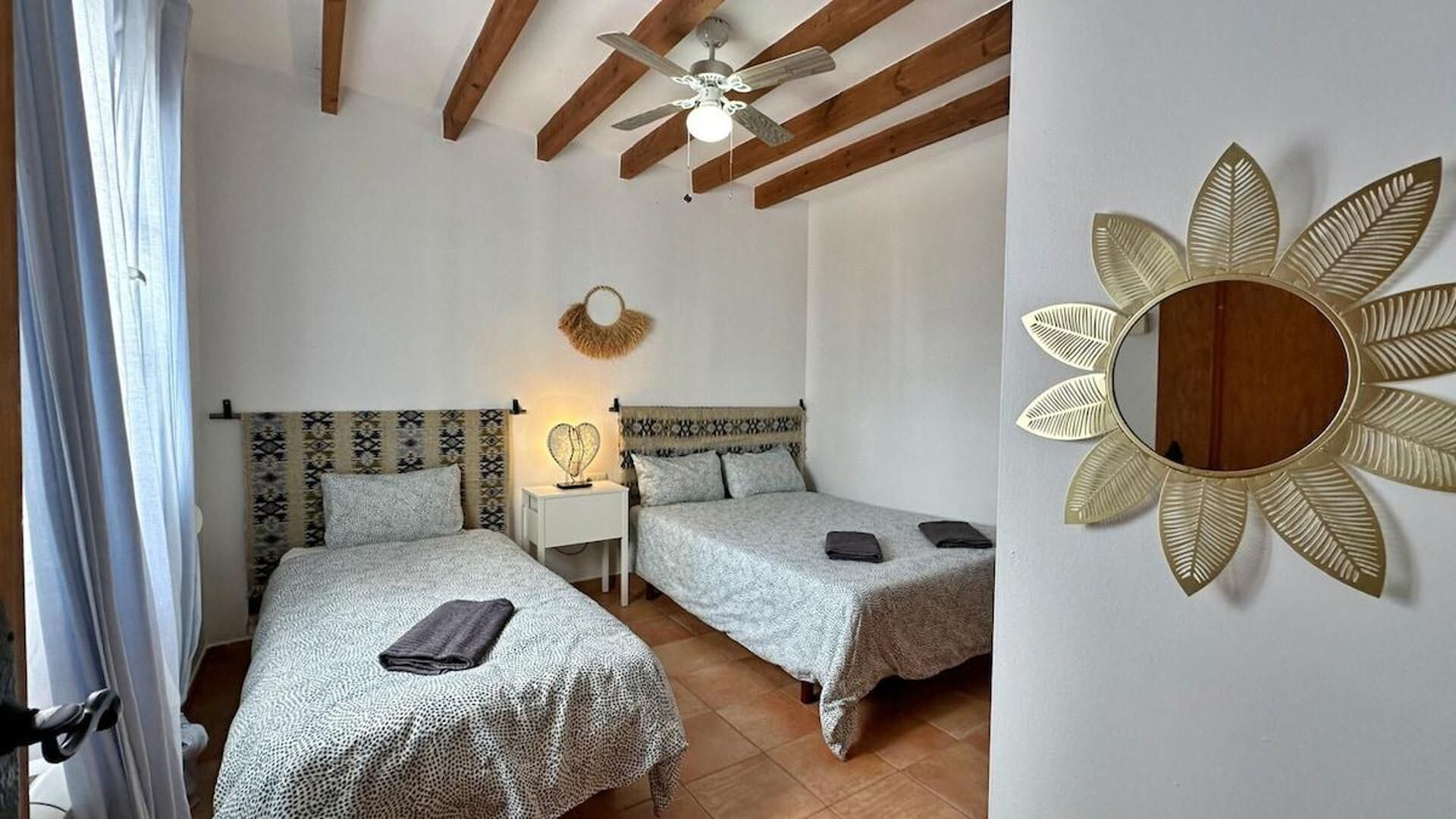 Una habitación con dos camas, ventilador de techo, espejo en la pared y una mesita con lámpara.