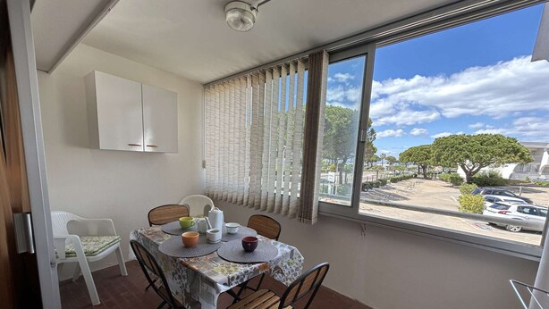 A balcony with a table set for two, a white cabinet, and a view of a parking lot and trees.