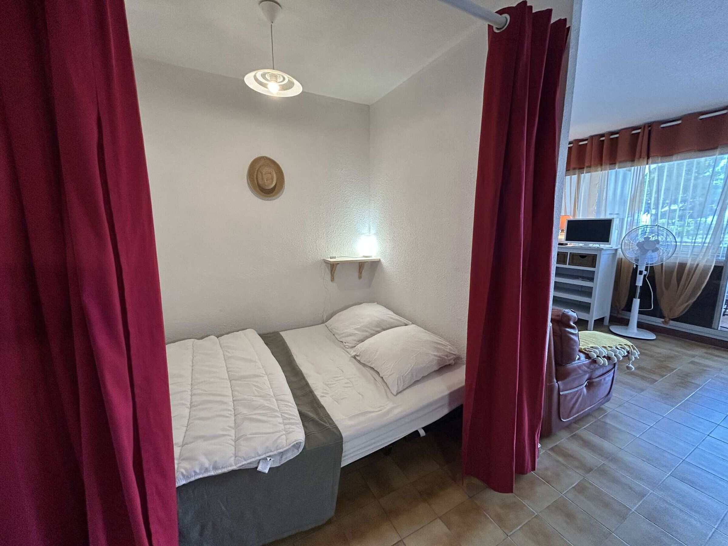 Une petite chambre à coucher, bien rangée, avec un lit, une table de chevet, une étagère fixée au mur, un ventilateur de plafond et une fenêtre avec des rideaux.