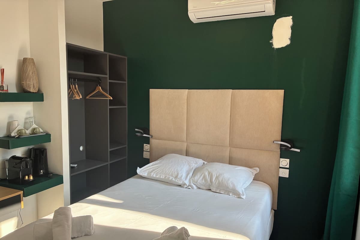 A neatly made bed with white linens and two pillows, a bedside table with a coffee maker, a hanging robe, and a wardrobe with shelves.
