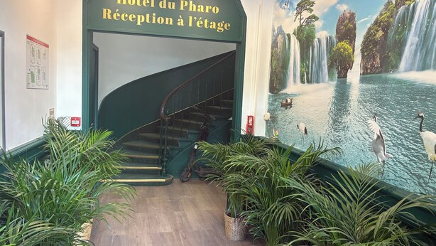 A hotel reception area with a staircase leading to the upper floor, a mural of a waterfall, and several potted plants. - hôtel du Pharo (Marseille)