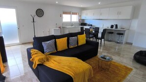 A living room with a black sofa, yellow throw, and patterned pillows. A dining area with a table and chairs is visible. A kitchen with white cabinets and a microwave is in the background. - The Infinite (Ribeira Grande)