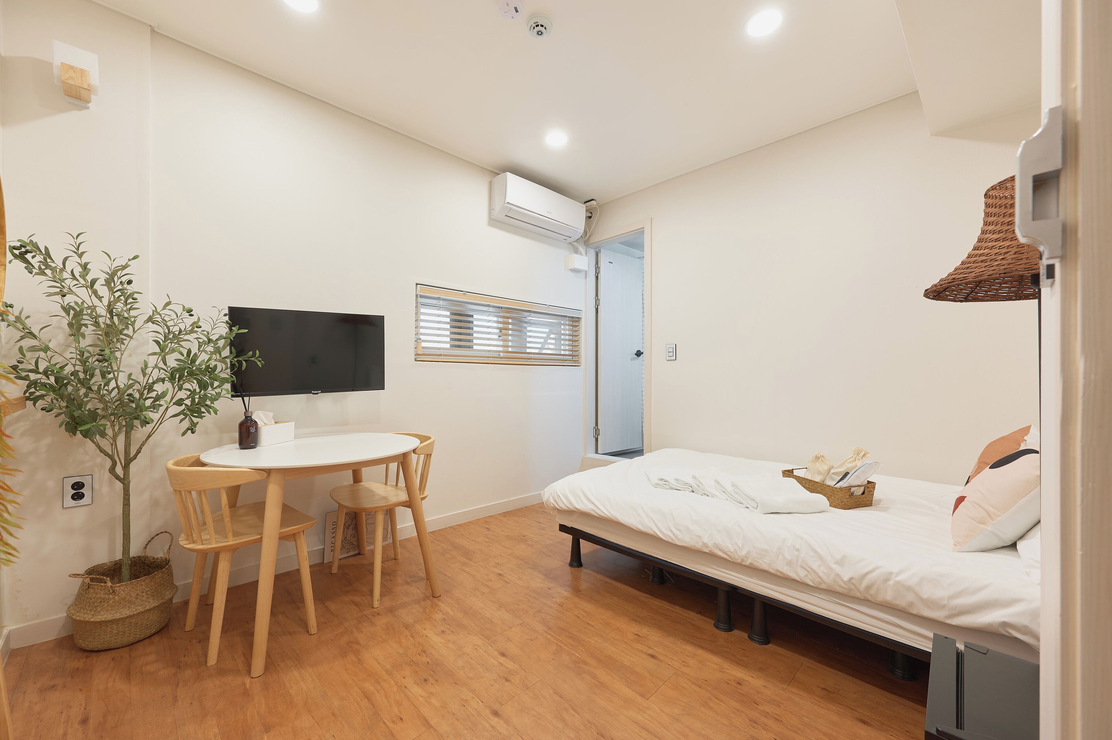 A small, clean room with a bed, a desk, a chair, a potted plant, and a TV.