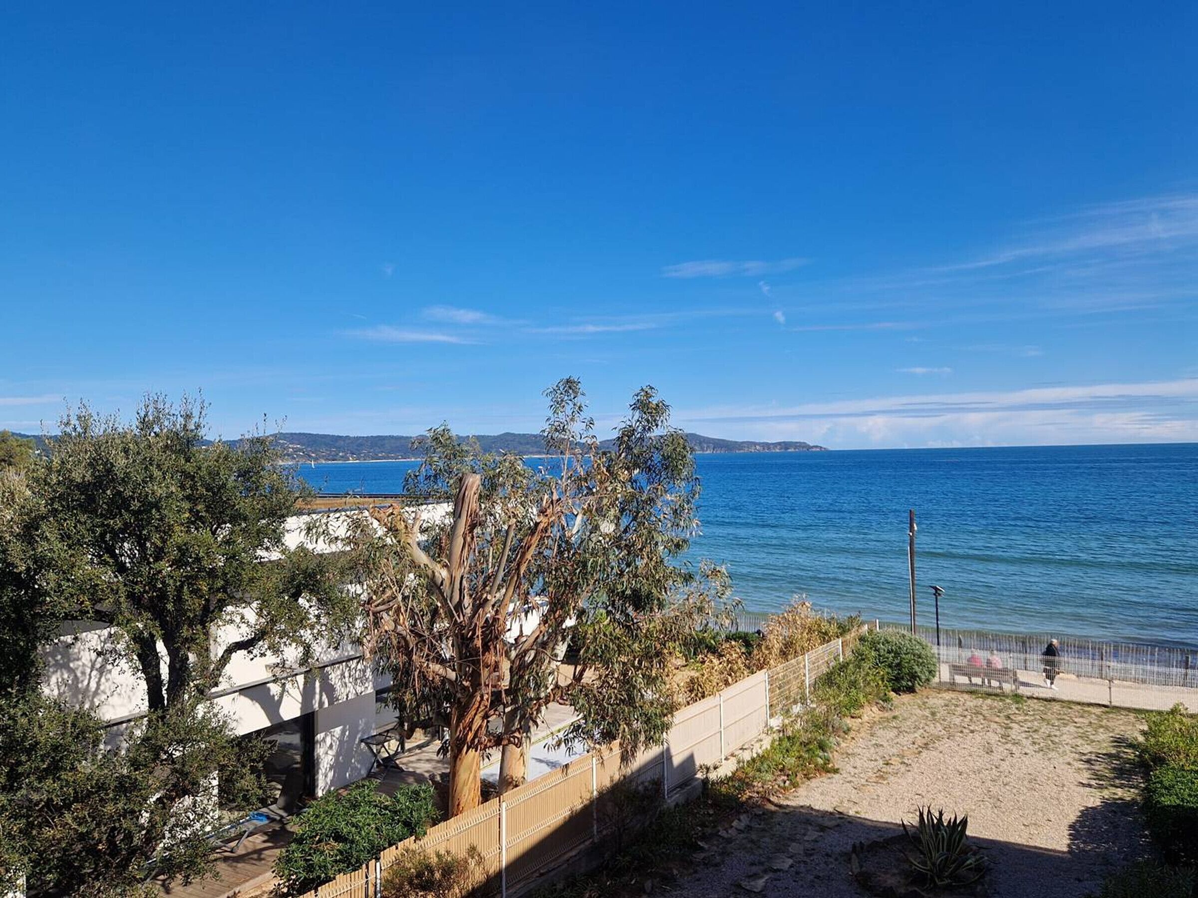 A coastal view with a sandy path, a fence, and trees in the foreground.