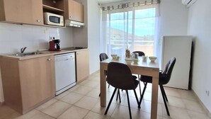 Eine kompakte Küche mit Mikrowelle, Herd und Geschirrspüler. Ein Esstisch mit Stühlen und ein Fenster mit Vorhängen sind vorhanden.