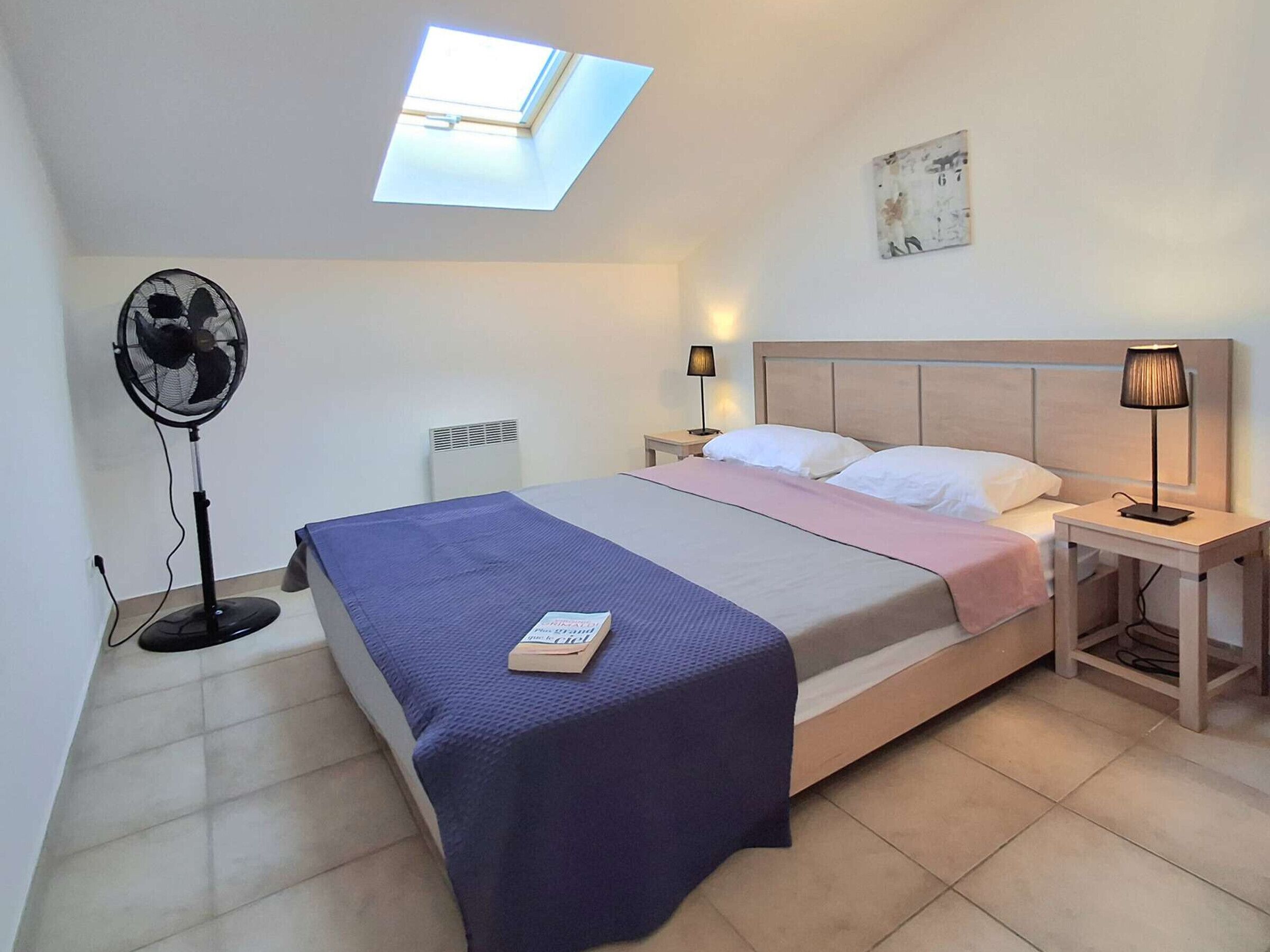 Een slaapkamer met een bed, een ventilator, een nachtkastje met een lamp, een radiator en een dakraam.
