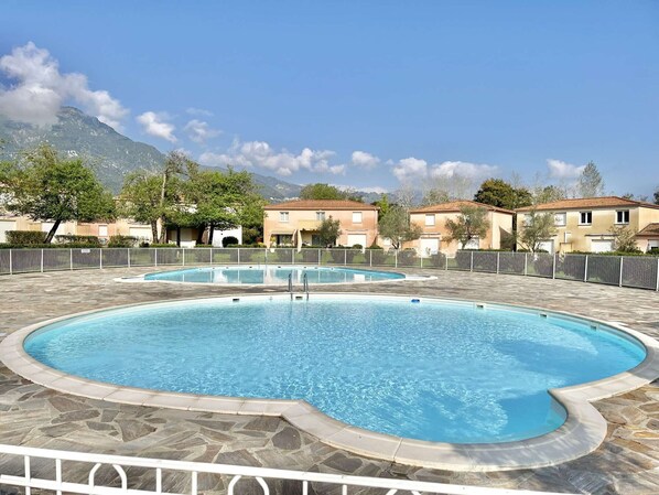 Una piscina con solárium de piedra, rodeada por una valla, y edificios residenciales al fondo.