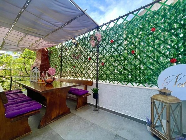 Un patio al aire libre con una mesa de madera, asientos acolchados morados, una chimenea de ladrillo y una pared de celosías verdes decoradas con flores.
