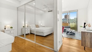 Quarto moderno com porta de vidro deslizante para uma varanda, cama com roupa de cama branca, mesa de cabeceira com abajur e uma mesinha lateral com uma planta.
