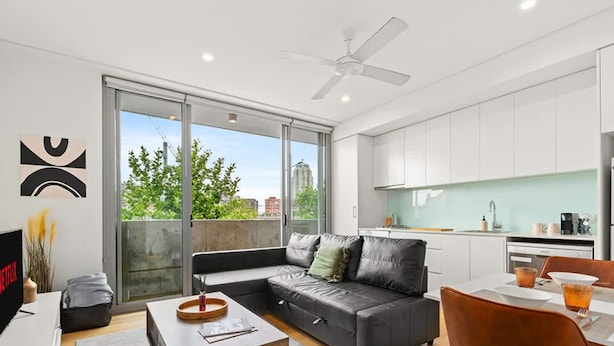 Un apartamento moderno con sala de estar, sofá, mesa de centro, televisor y área de cocina con vista al exterior.