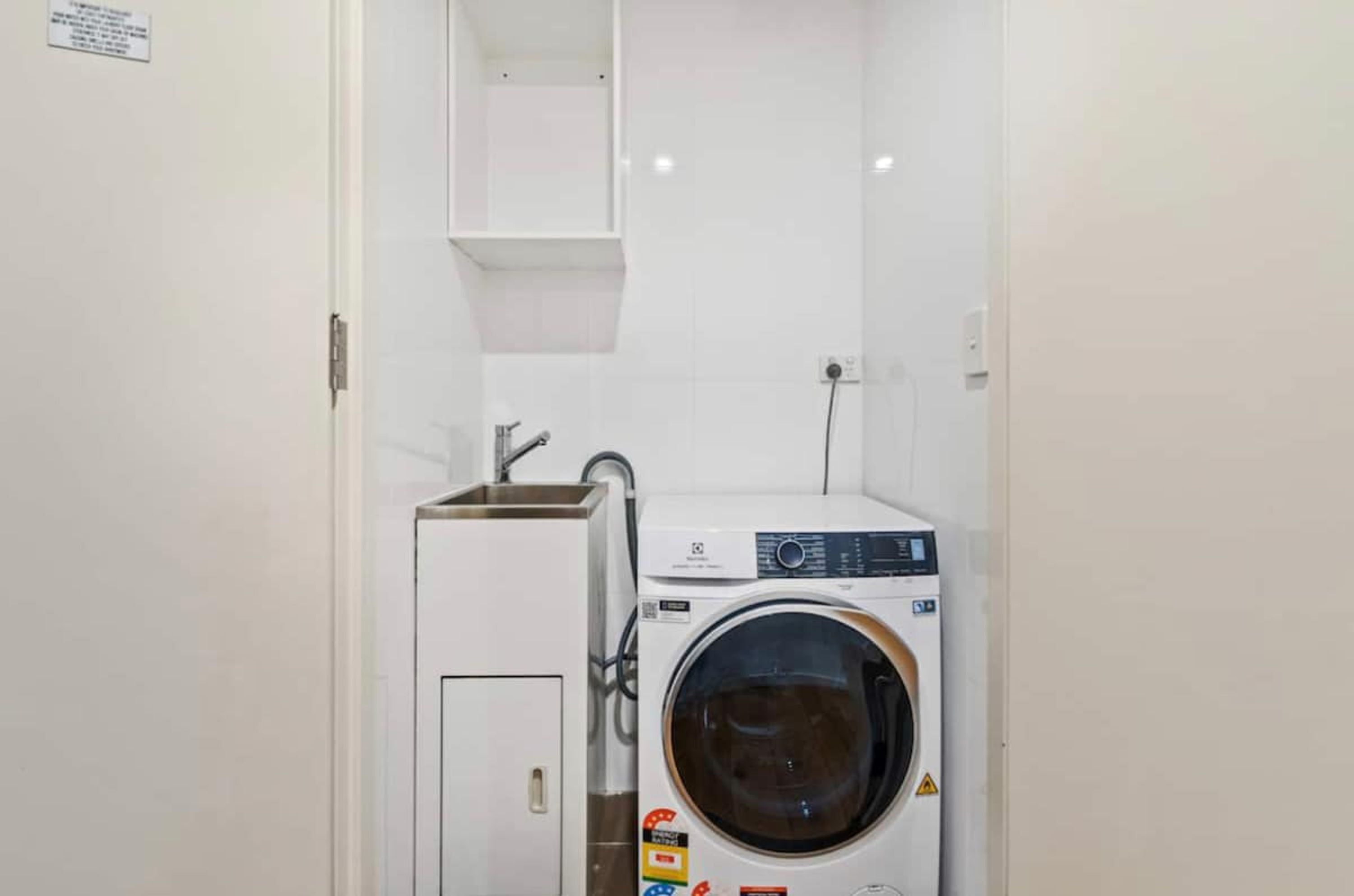 Une petite buanderie comprenant un lave-linge et un sèche-linge, une petite étagère au-dessus du sèche-linge et une porte avec une pancarte.