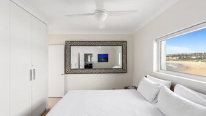 A bedroom with a bed, a ceiling fan, a large window with a beach view, and a mirror with a black frame. - Beachfront Luxury 3 Beds With Ocean View Balcony (Sydney)
