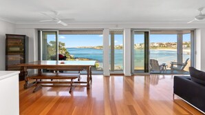 A spacious room with a wooden floor, a dining table, a sofa, and large sliding glass doors leading to a balcony with a view of the sea. - Beachfront Luxury 3 Beds With Ocean View Balcony (Sydney)