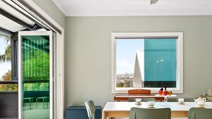 Un coin repas avec une table dressée pour deux personnes, une vue sur le paysage urbain par une fenêtre et une porte-fenêtre coulissante donnant sur un espace extérieur.