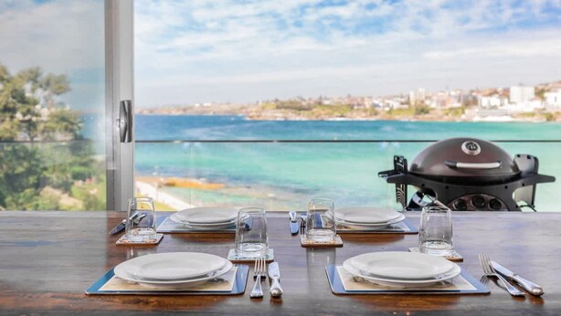 Mesa de comedor para cuatro con vista al mar y una parrilla al fondo.