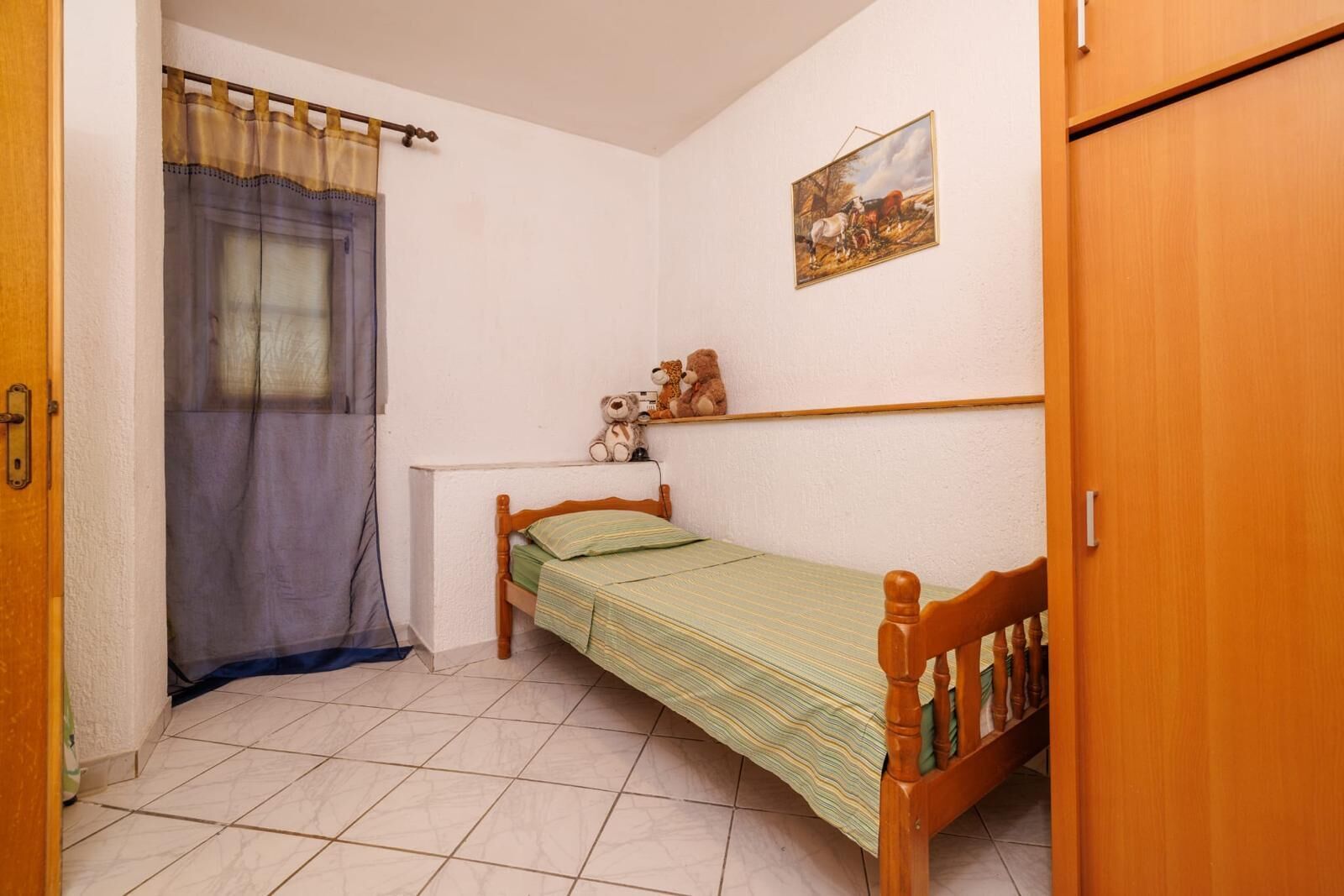 A small, single-bed room with a wooden headboard, a window with a curtain, a painting on the wall, and a door.