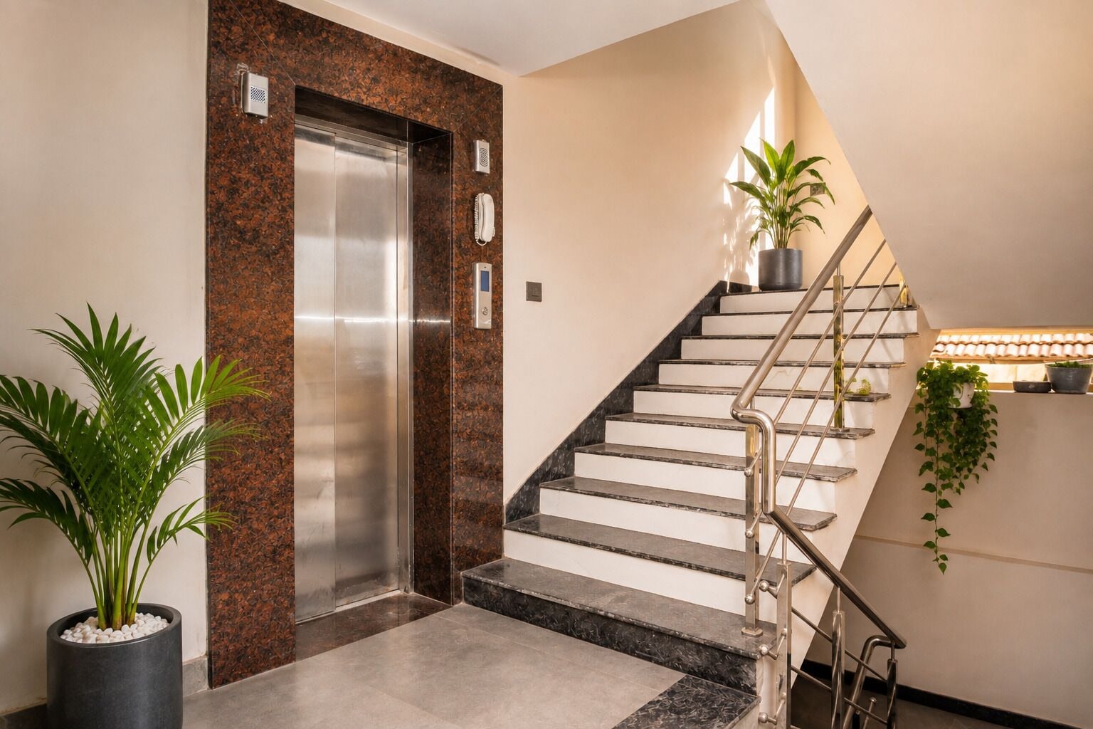 Un ascensor moderno con detalles metálicos, una escalera con barandilla de metal y plantas en macetas.