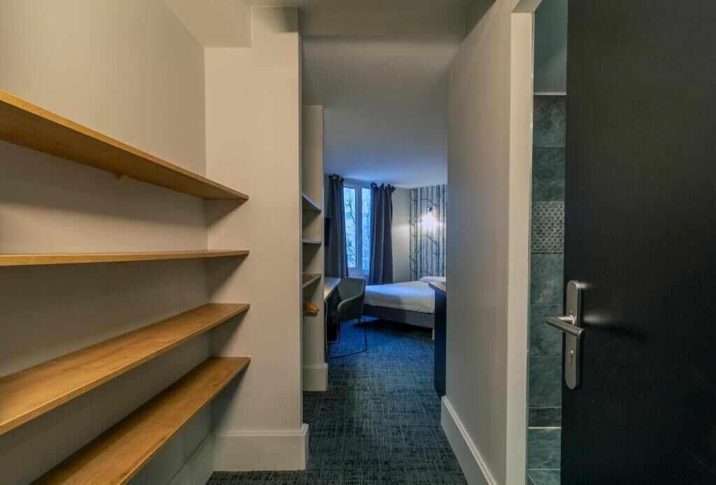 A narrow hallway with wooden shelves, a bed with a blue headboard, and a door with a handle.