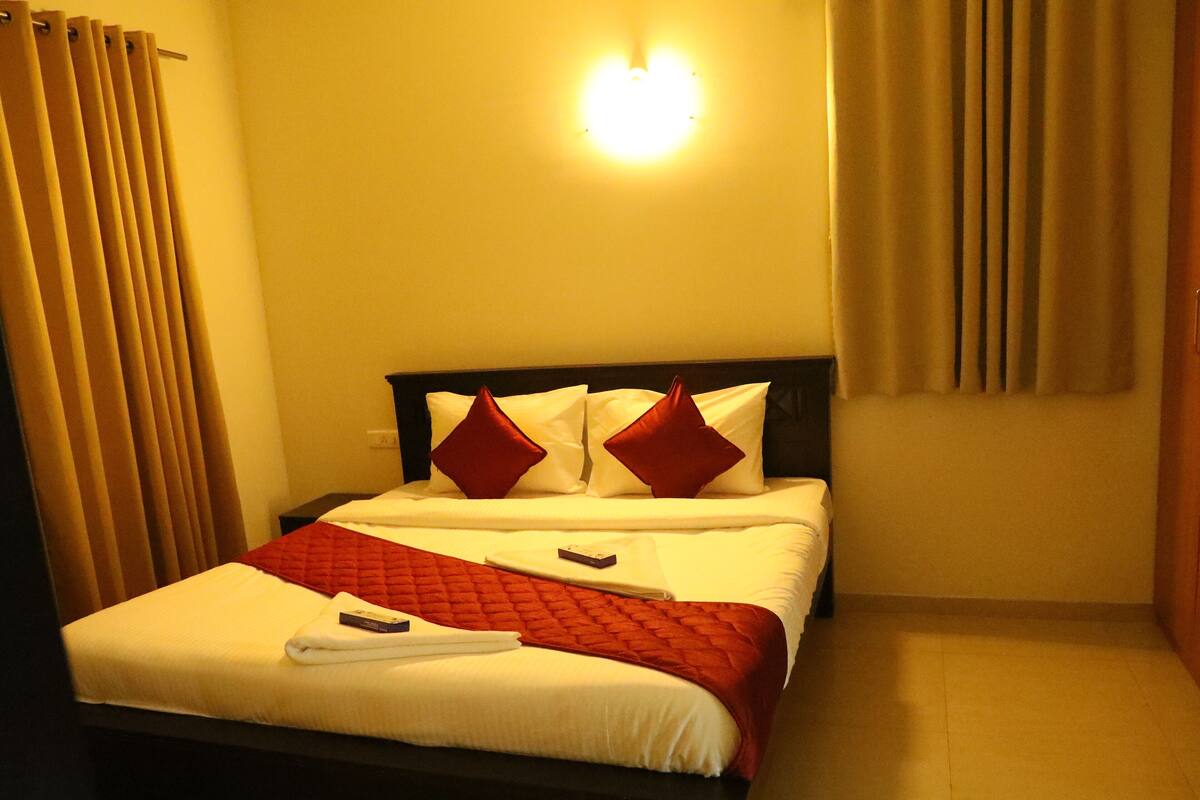 A hotel room with a bed, curtains, a wall-mounted light, and a remote control on the bed.
