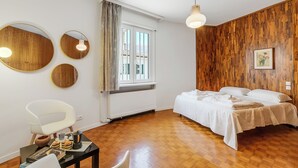 Una habitación con suelo de madera, una cama con ropa de cama blanca, un espejo redondo, una silla blanca y una mesa con libros y una taza.