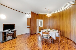 Una habitación con piso de madera, una mesa de comedor con sillas, un televisor sobre un mueble bajo y una puerta que da a otra habitación.