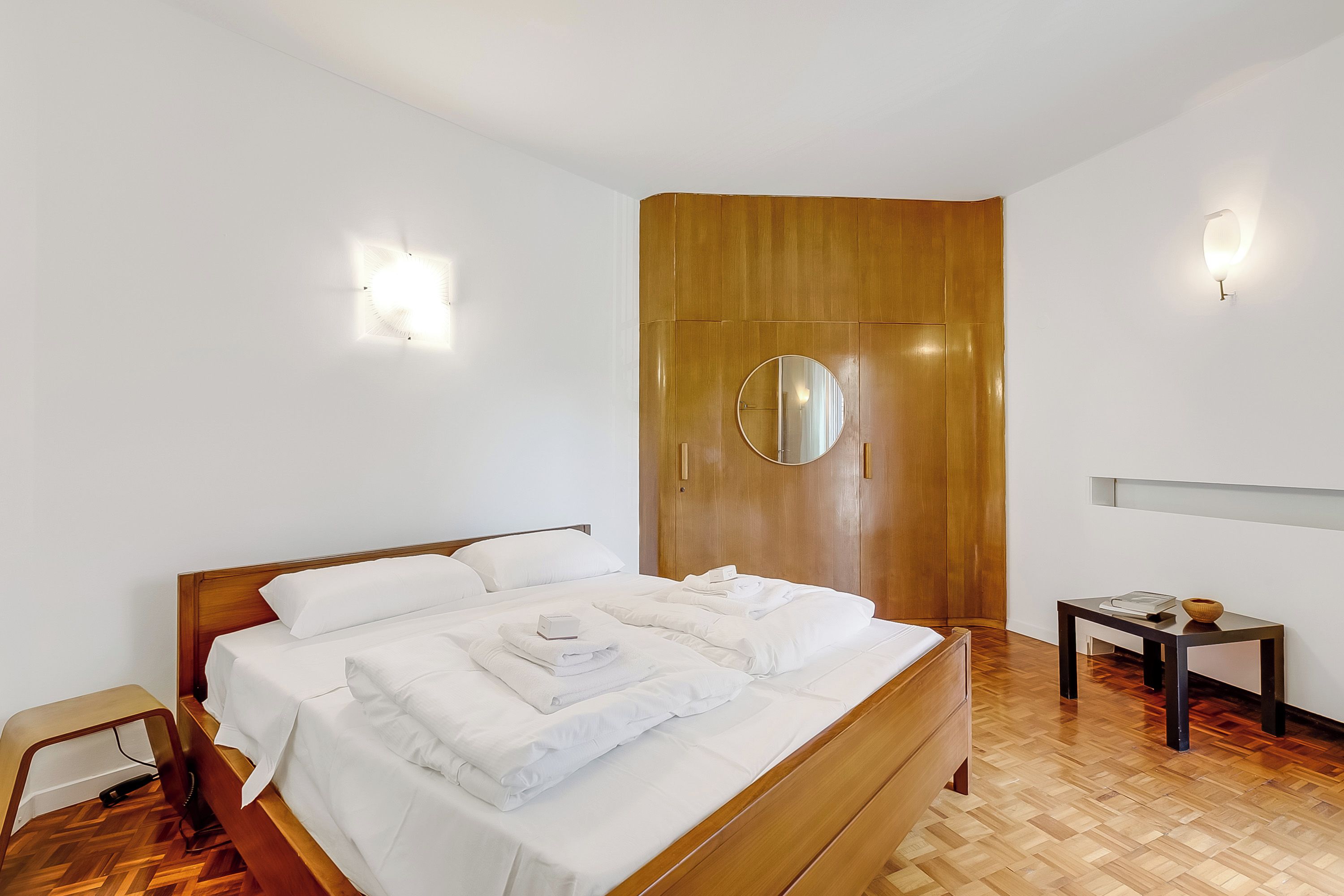 Een netjes opgemaakt bed met witte lakens, een houten garderobe, een klein tafeltje met een schaal en een wandspiegel.