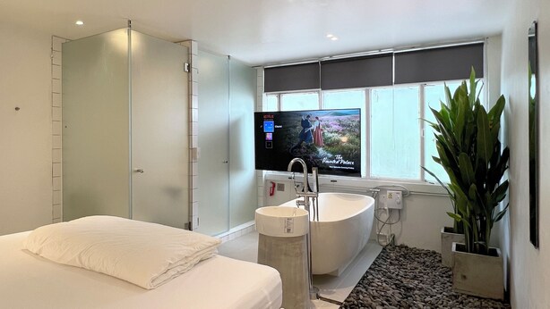 A modern bedroom with a large bed, a built-in TV, a bathtub, and a large window. - Chatel Samyan BKK (Bangkok)