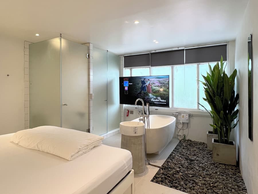 A modern bedroom with a large bed, a built-in TV, a bathtub, and a large window.