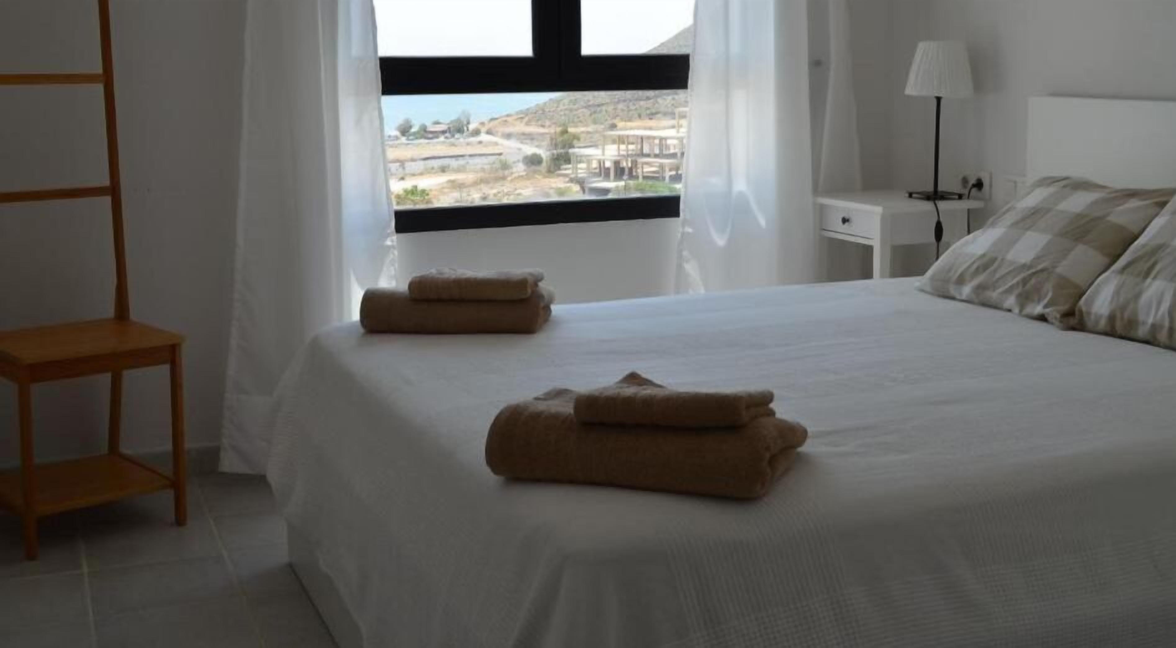 A neatly made bed with two rolled towels, a bedside table with a lamp, and a window with a view of buildings and hills.