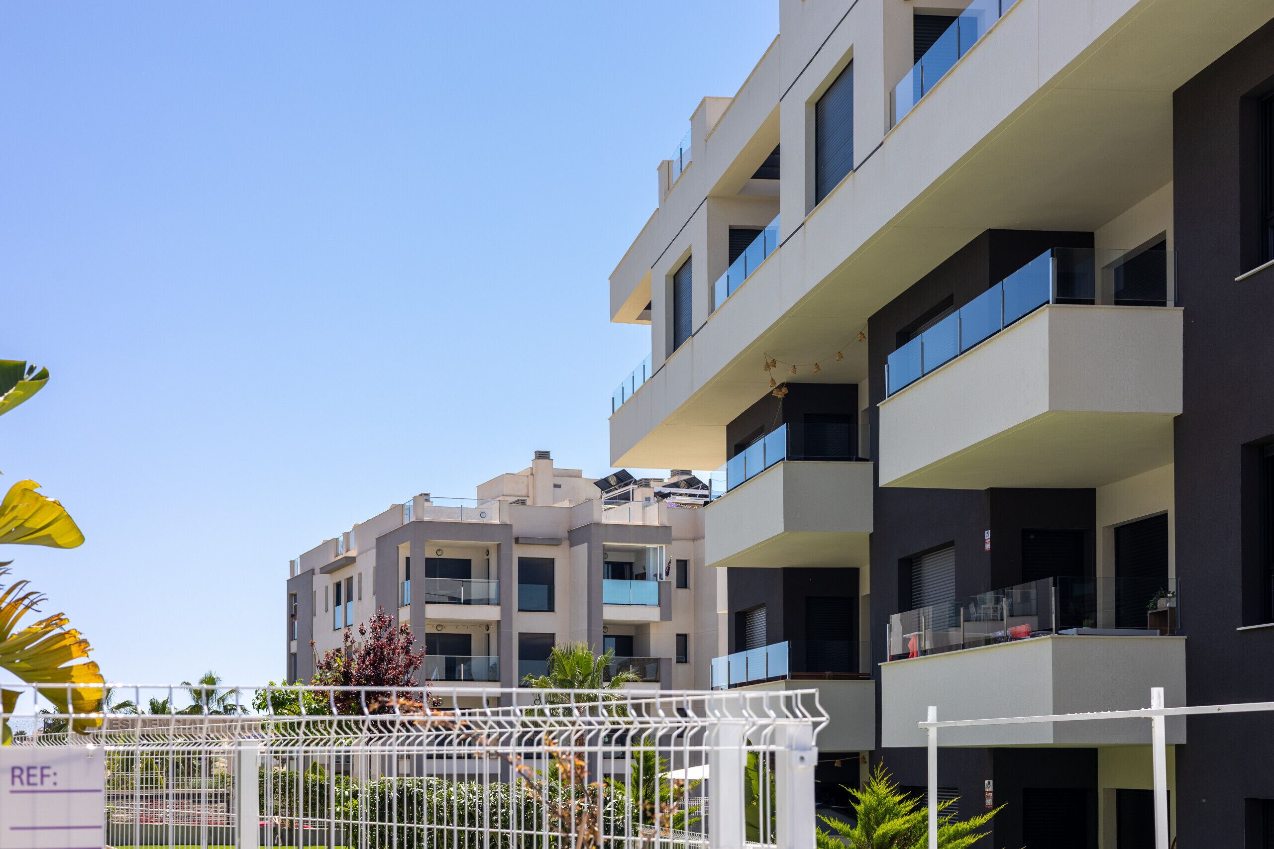A modern multi-story residential building with balconies, a gated entrance, and a clear sky.