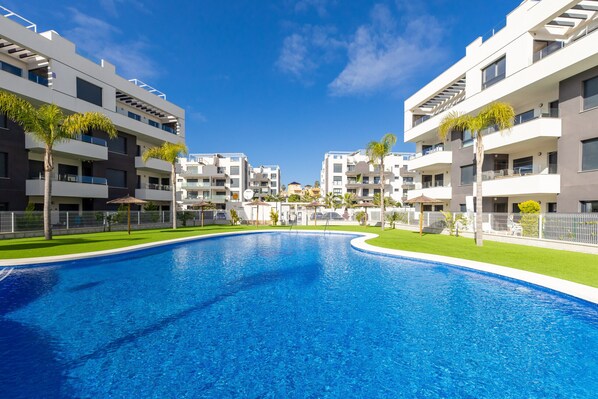 A modern pool area with a clear blue pool, surrounded by multi-story buildings with balconies and palm trees.