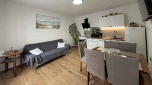 Un espace de vie compact comprenant un canapé, une table à manger, des chaises, un coin cuisine avec une cuisinière, un évier et des placards, ainsi qu’un téléviseur fixé au mur.