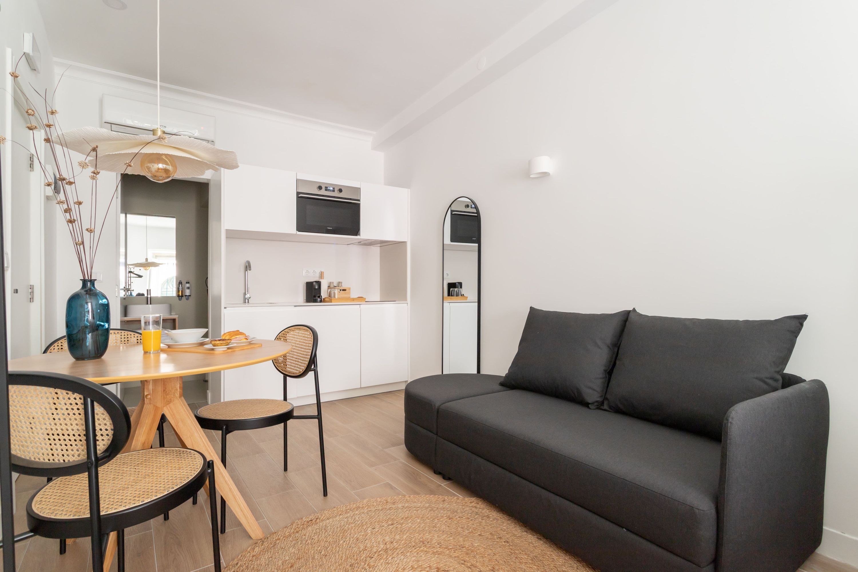 A modern living room with a dark sofa, a dining table with chairs, a kitchen area with white cabinets, and a large mirror on the wall.