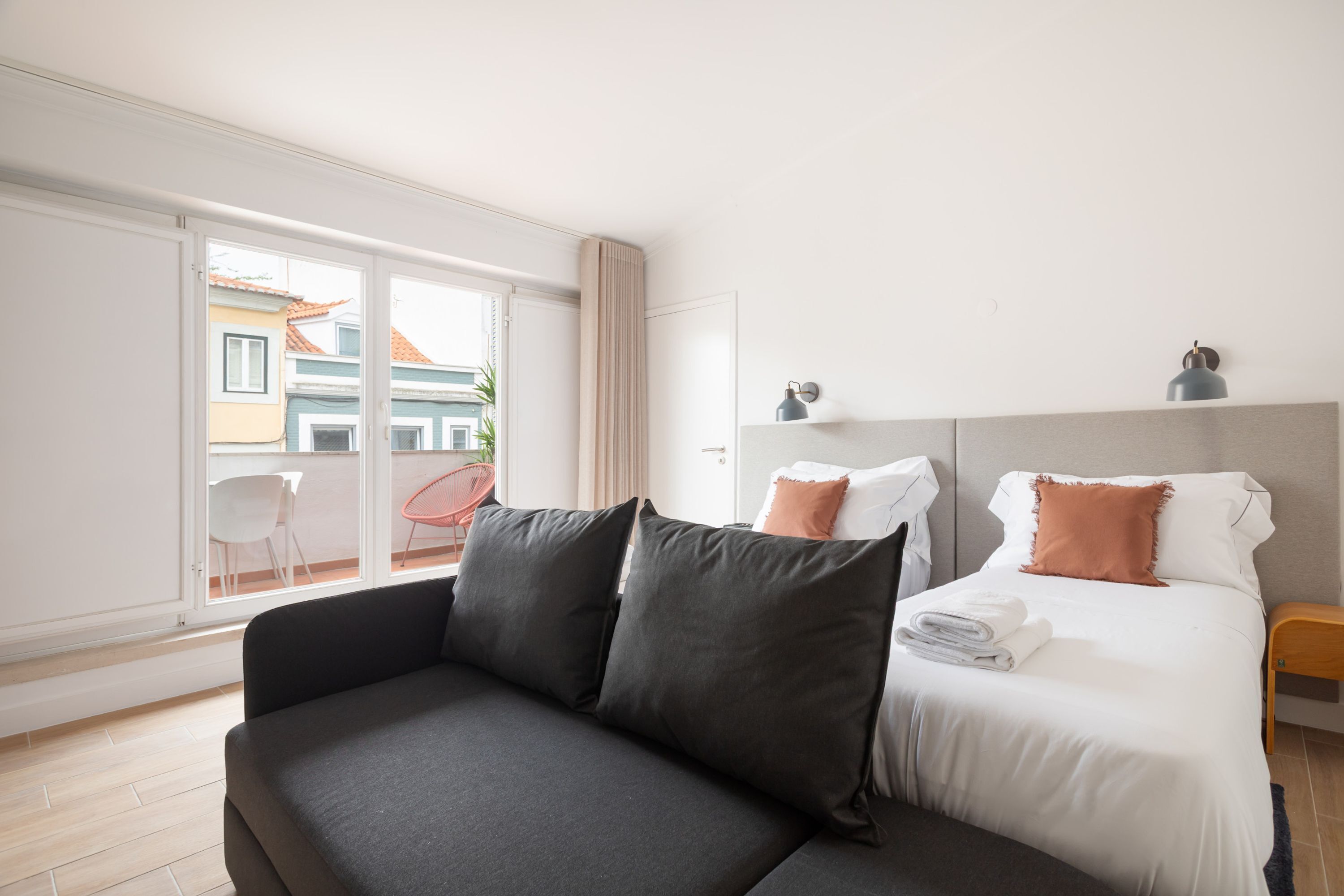 A modern hotel room with a sofa, a neatly made bed, a small bedside table, and a view through a large window.