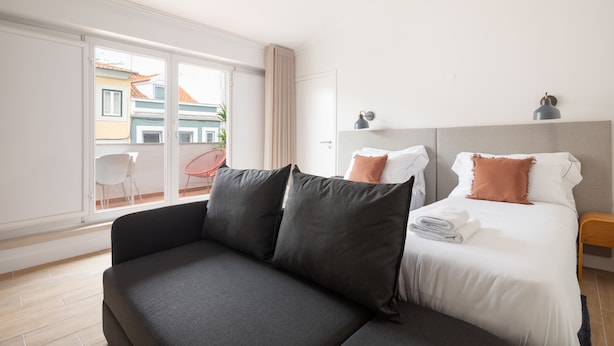 A modern hotel room with a sofa, a neatly made bed, a small bedside table, and a view through a large window. - Lapa Diplomatic Flat 1 1 by Feel Lisboa (Lisbon)